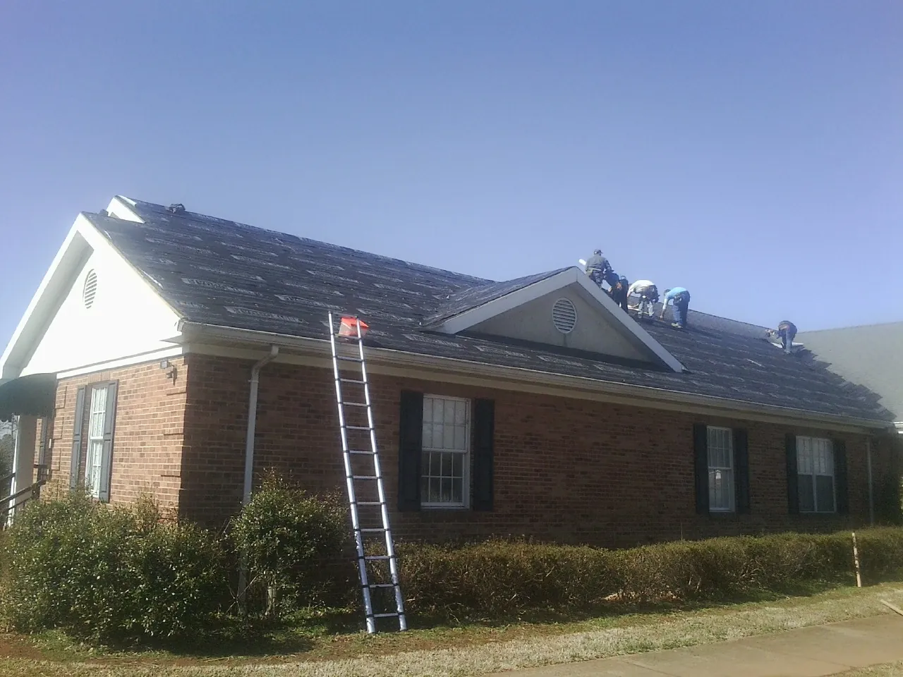 Crew tearing shingles off a brick ranch roof with a ladder set — Emergency Roof Repair work by our crew in Clarence